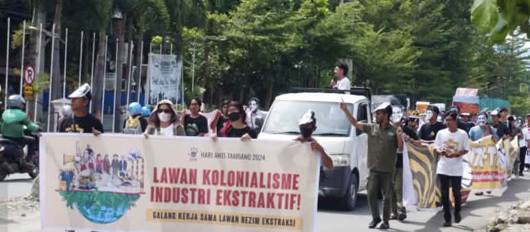 Dengan berbagai aksi, teater, musik dan pameran, para penjaga lingkungan dan warga yang terkena dampak menentang perusakan oleh pertambangan Demo – tampak depan: spanduk “Lawan Kolonialisme Industri Ekstraktif”, tampak belakang: peserta demo dan sebuah mobil pick-up