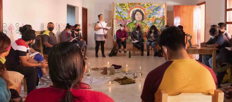 Pelatihan tentang keadilan energi di Chiapas, pemikiran mencari alternatif bagi kedaulatan di Puebla, Meksiko Para peserta duduk berlingkar. Mereka mengikuti sebuah workshop. Di tengah-tengah berdiri lilin yang menyala dan tergeletak kertas yang bertuliskan PAZ. Di dinding tergantung sebuah lukisan wajah Berta Cáceres.