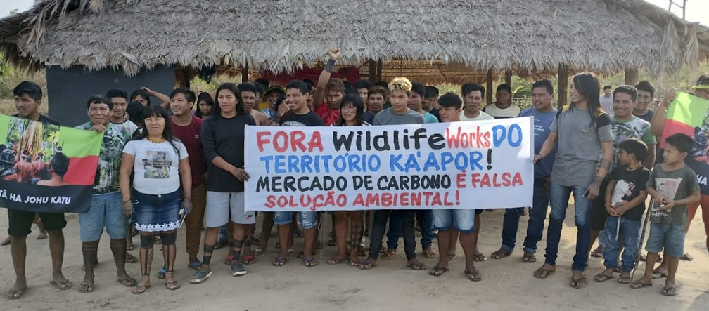 Suku Ka’apor protes menentang bisnis dengan hutan hujan mereka. Tulisan spanduk: „Wildlife Works keluar dari wilayah Ka’apor. Perdagangan karbon adalah pemecahan krisis lingkungan yang keliru.“ Sebuah kelompok terdiri dari 30 anggota masyarakat adat di depan pondok yang beratapkan pelepah kelapa melakukan protes dengan memegang spanduk: "Wildlife Works keluar dari wilayah Ka’apor. Perdagangan karbon adalah pemecahan krisis lingkungan"