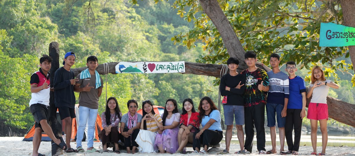 Sebuah kelompok anak muda sedang duduk dan berdiri di depan pohon di pantai