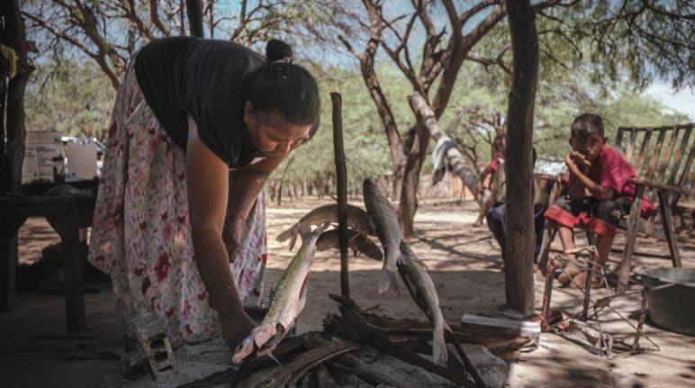 Seorang perempuan adat di bawah pepohonan sedang bersiap memanggang ikan, sementara seorang anak menatapnya.